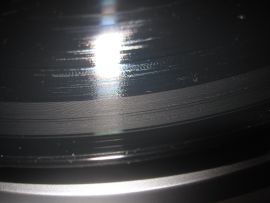 Close-up photo shows a black record with a gray ring starting from the outer edge and continuing through the first track. Damage was from a defective stylus on a Grace Digital Audio Vinylwriter (AVPUSB01S) USB turntable.